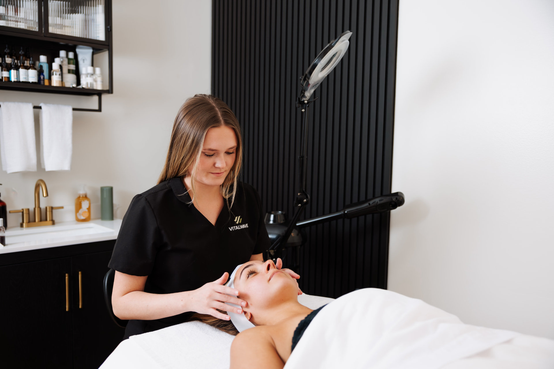 A skincare therapist in a black uniform gives a facial to a client lying on a treatment bed